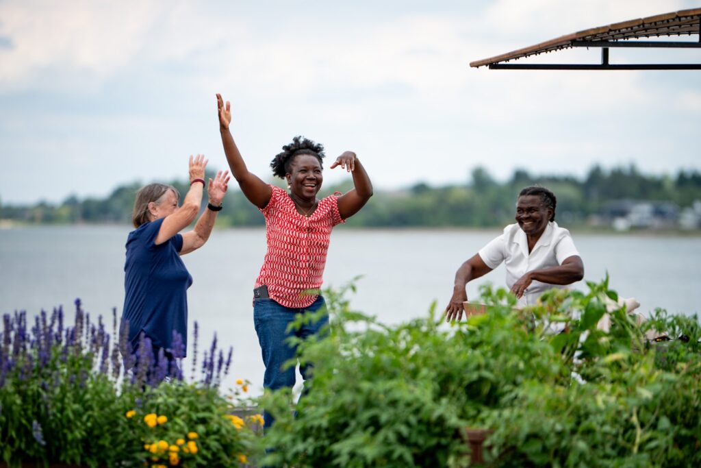 Trois femmes souriantes partagent un moment de joie et de complicité dans un jardin communautaire au bord de l’eau, illustrant l’inclusion et la convivialité en milieu régional.